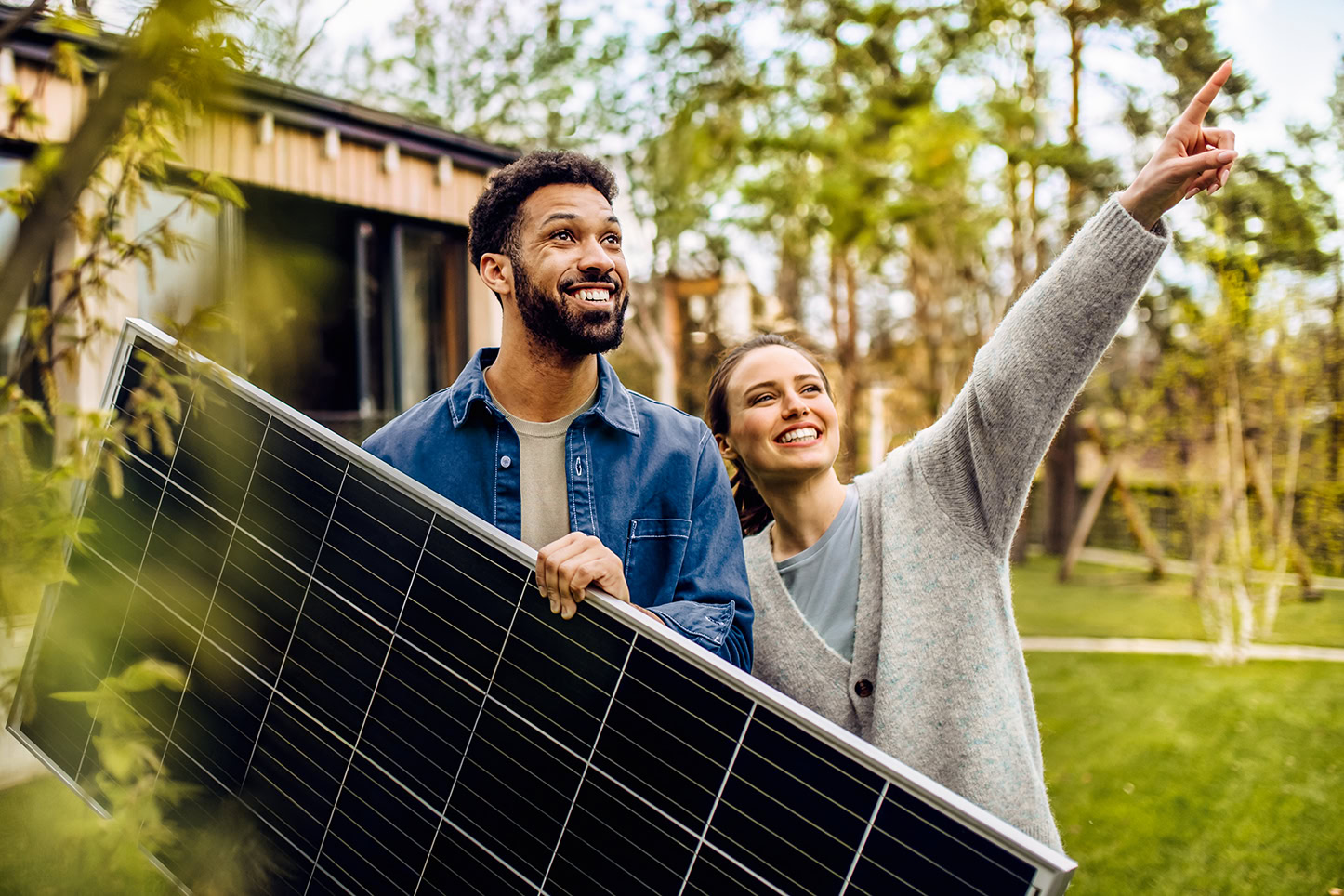 Ein lächelnder Mann hält ein Solarpanel, während eine Frau neben ihm nach oben zeigt. Sie stehen draußen in einem grünen Garten mit Bäumen und einem Haus im Hintergrund, was auf einen umweltfreundlichen oder nachhaltigen Lebensstil hindeutet.