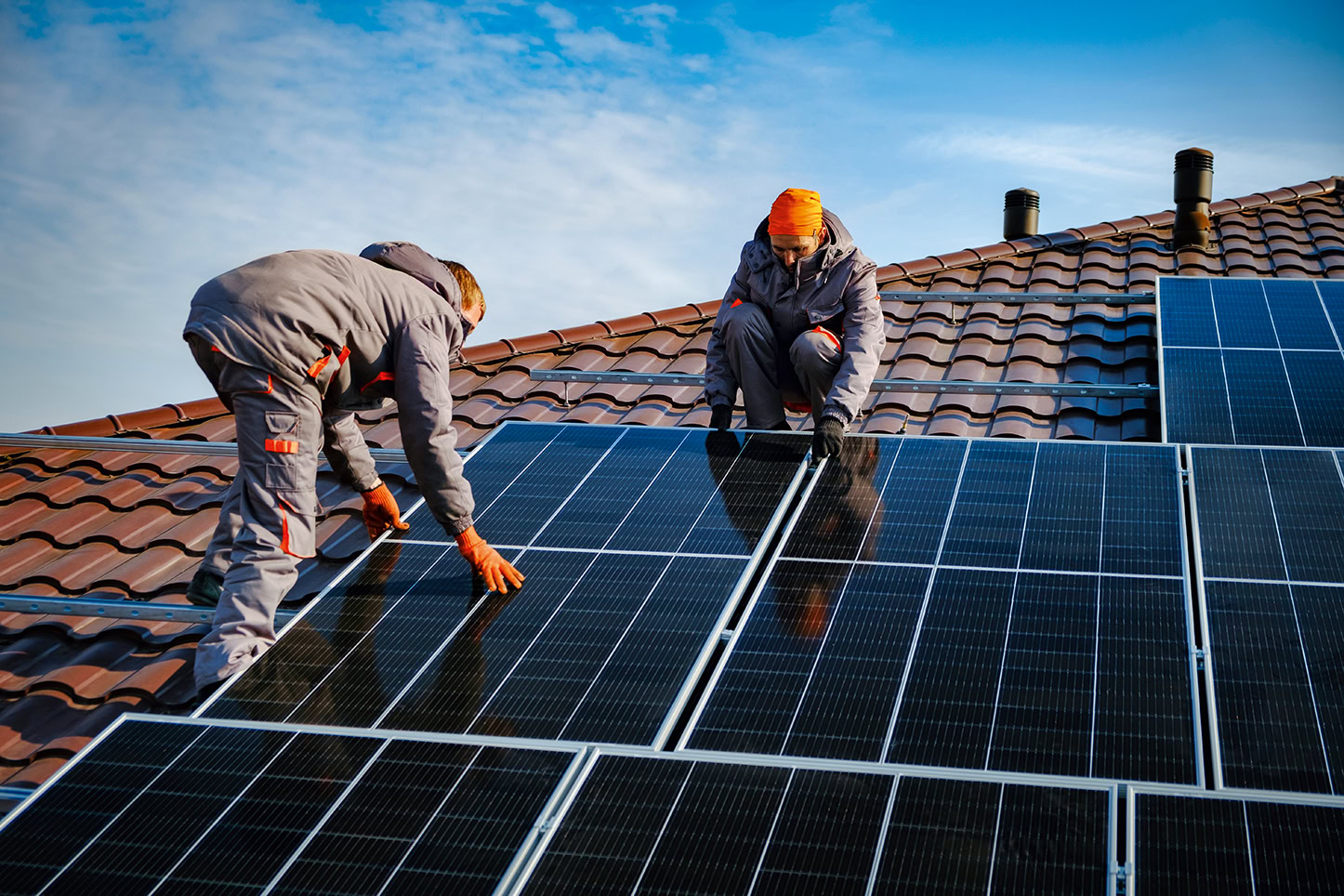Zwei Arbeiter in grauen Uniformen und orangefarbenen Handschuhen installieren unter einem blauen Himmel mit leichten Wolken Solarzellen auf einem braunen Ziegeldach.