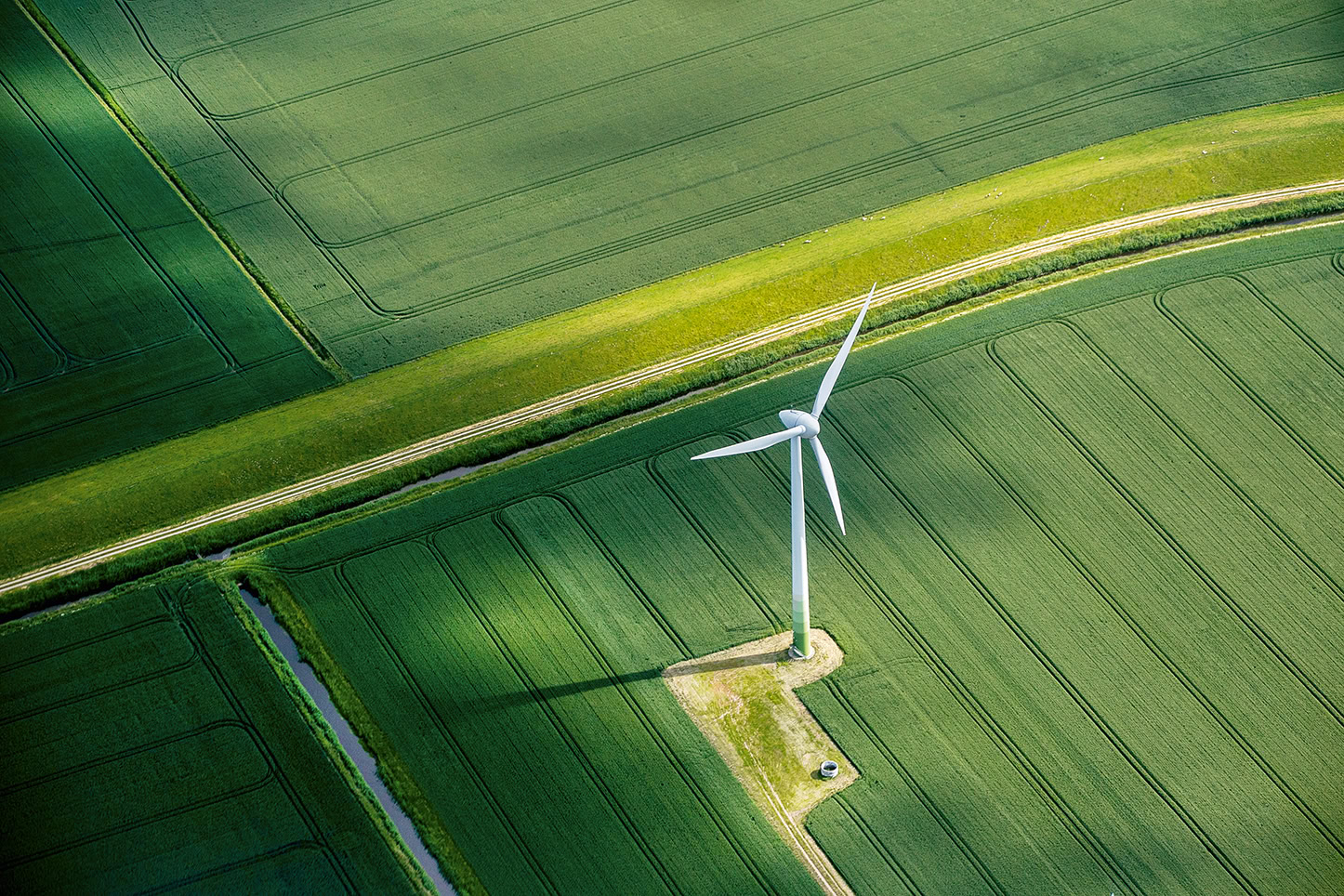 Luftaufnahme einer einzelnen Windkraftanlage, die inmitten grüner landwirtschaftlicher Felder mit sich kreuzenden Wegen steht und einen langen Schatten auf die darunter liegenden Pflanzen wirft.