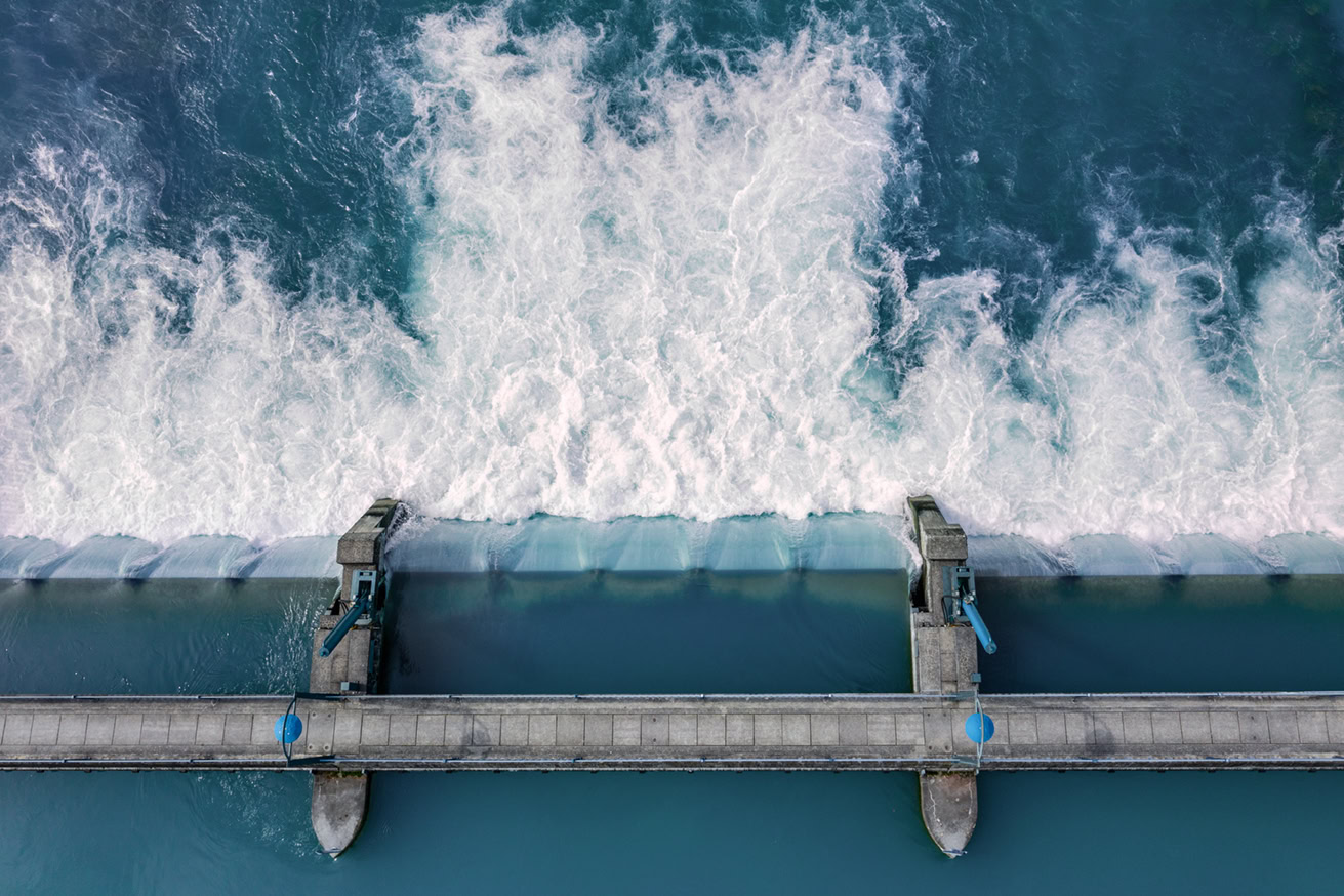 Luftaufnahme eines Staudamms mit Wasser, das über die Überlaufrinne fließt und weiße, schäumende Wellen erzeugt. Ein Steg mit Geländern und blauen Maschinen überquert die Dammstruktur.