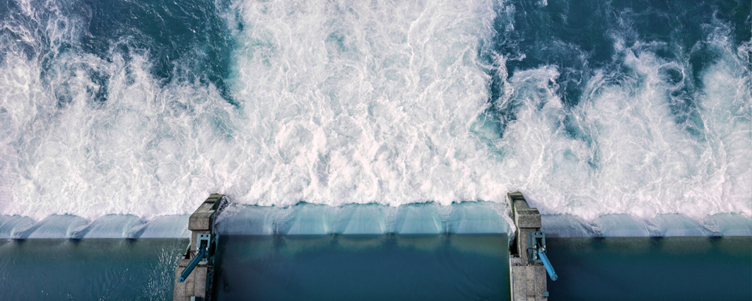 Luftaufnahme von Wasser, das mit großer Wucht über einen Überlauf eines Staudamms fließt, mit weißen, schäumenden Wellen, die unter ihm zusammenschlagen, und mechanischen Strukturen auf beiden Seiten.