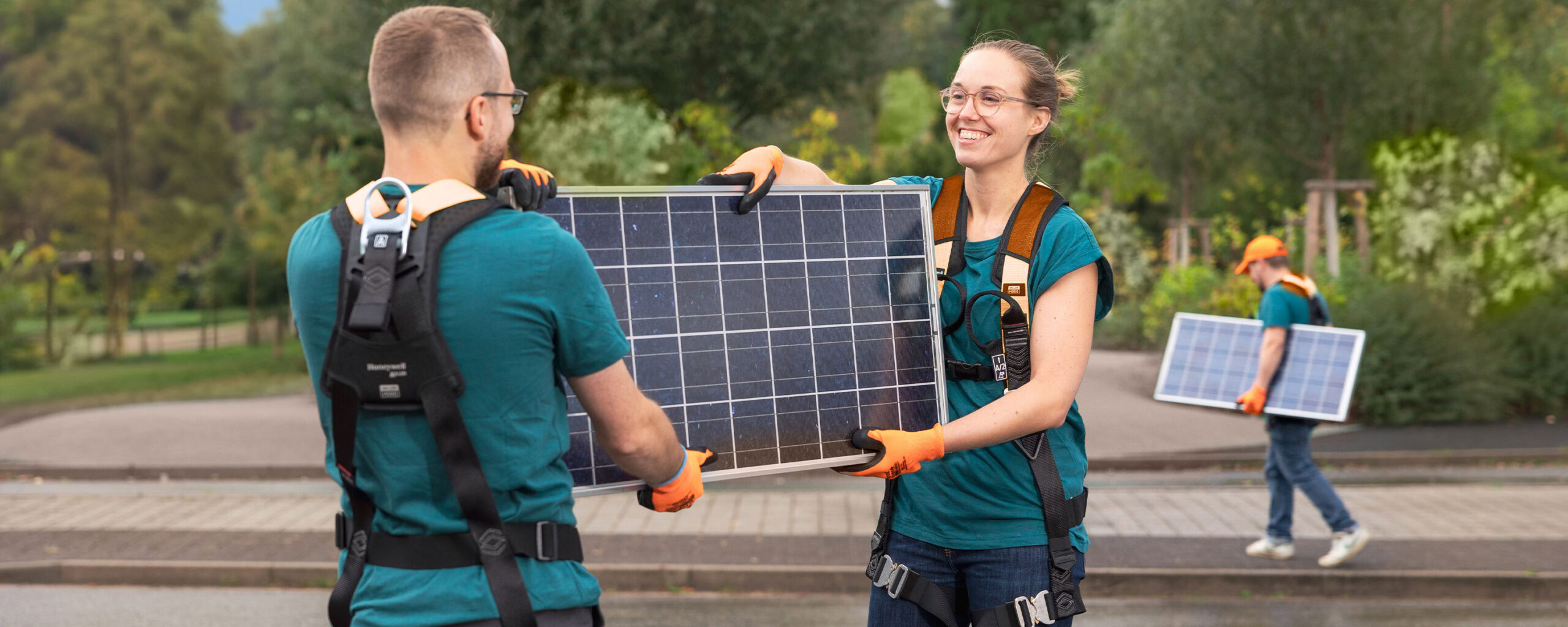 Zwei Personen mit Sicherheitsgurten und Handschuhen tragen lächelnd ein Solarmodul im Freien. Im Hintergrund trägt eine weitere Person ein Solarmodul. Bäume und Grünflächen umgeben die Szene.
